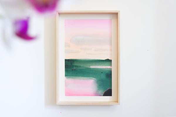 A framed abstract landscape original painting featuring soft pink sky, white clouds and teal watercolour washes of green field, mounted on a white wall with a light wood frame.