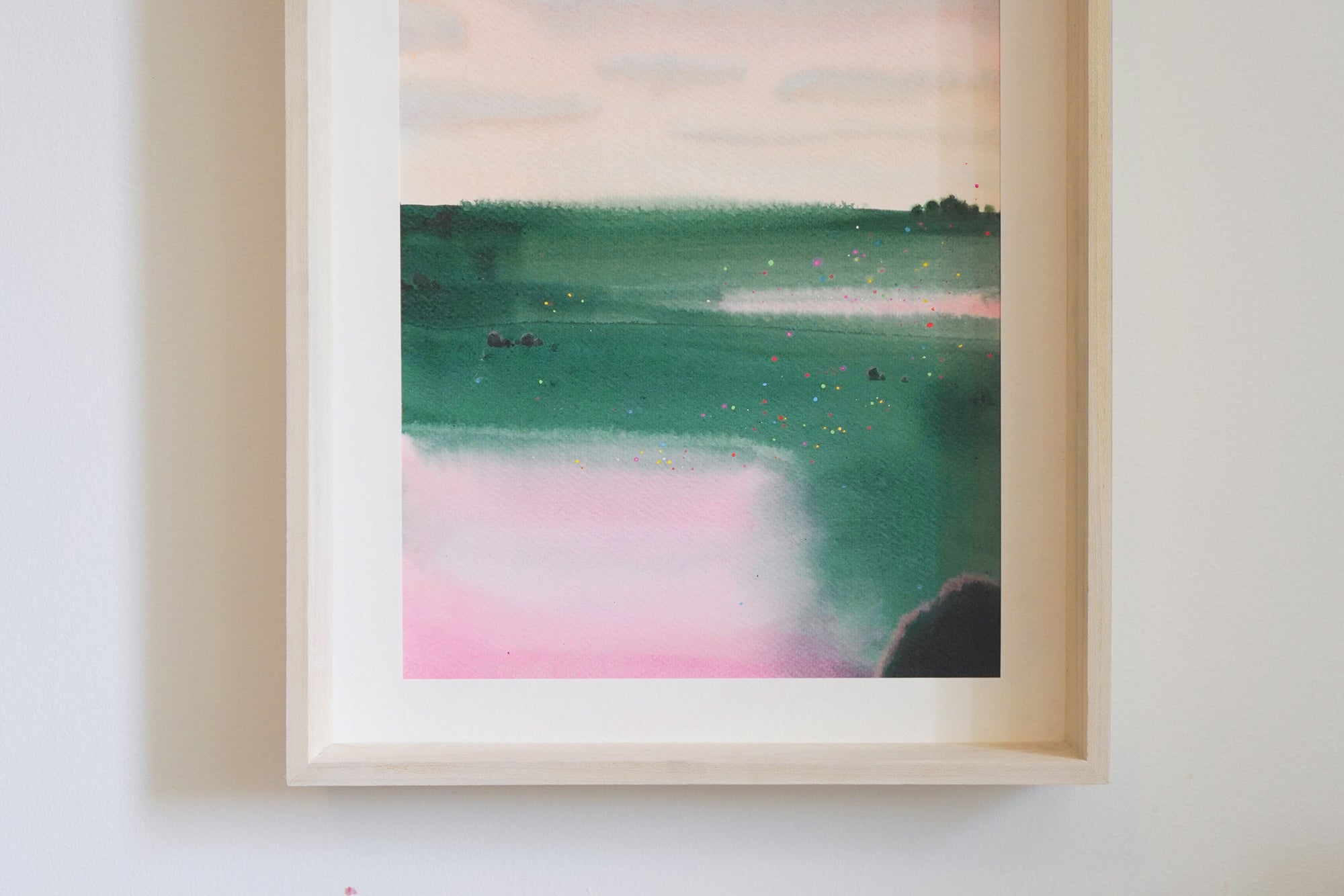A framed abstract landscape painting featuring shades of green, teal, pink, and cream, scene of a summer field mounted on a light-colored wall.