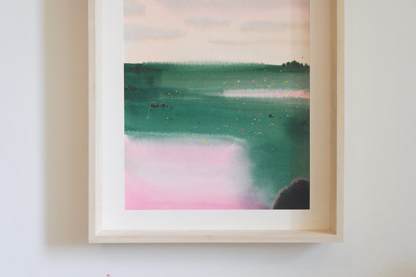 A framed abstract landscape painting featuring shades of green, teal, pink, and cream, scene of a summer field mounted on a light-colored wall.