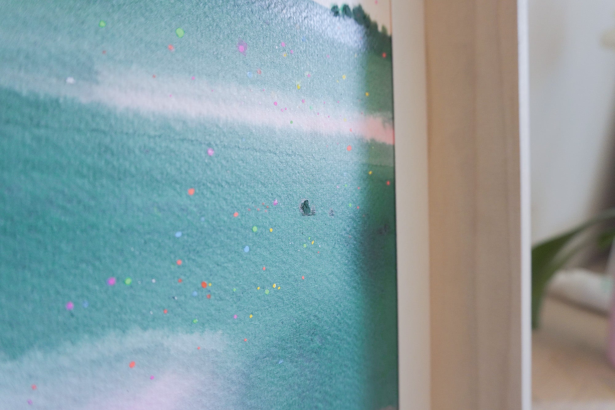 A close-up of a framed artwork showing a textured teal and turquoise landscape with small scattered colorful dots, photographed indoors against a light neutral background.