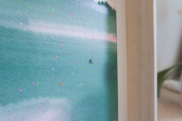 A close-up of a framed artwork showing a textured teal and turquoise landscape with small scattered colorful dots, photographed indoors against a light neutral background.
