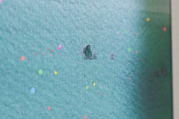 A close-up of a textured oil pastel teal-green painting surface with scattered small multicolored confetti dots and a small dark hole or stain near the center.