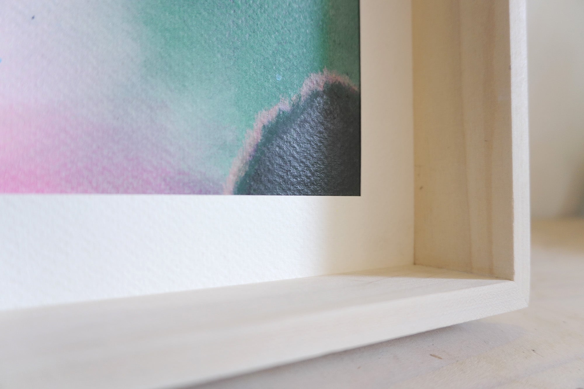 Close-up of a framed abstract original painting  showing a textured watercolor design with teal, pink, and dark gray tones inside a light wood frame.