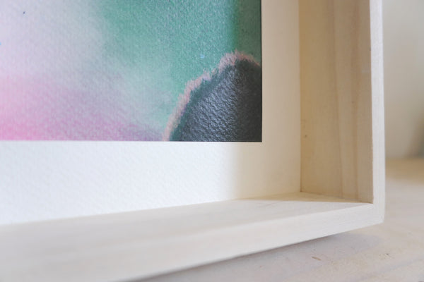 Close-up of a framed abstract original painting  showing a textured watercolor design with teal, pink, and dark gray tones inside a light wood frame.