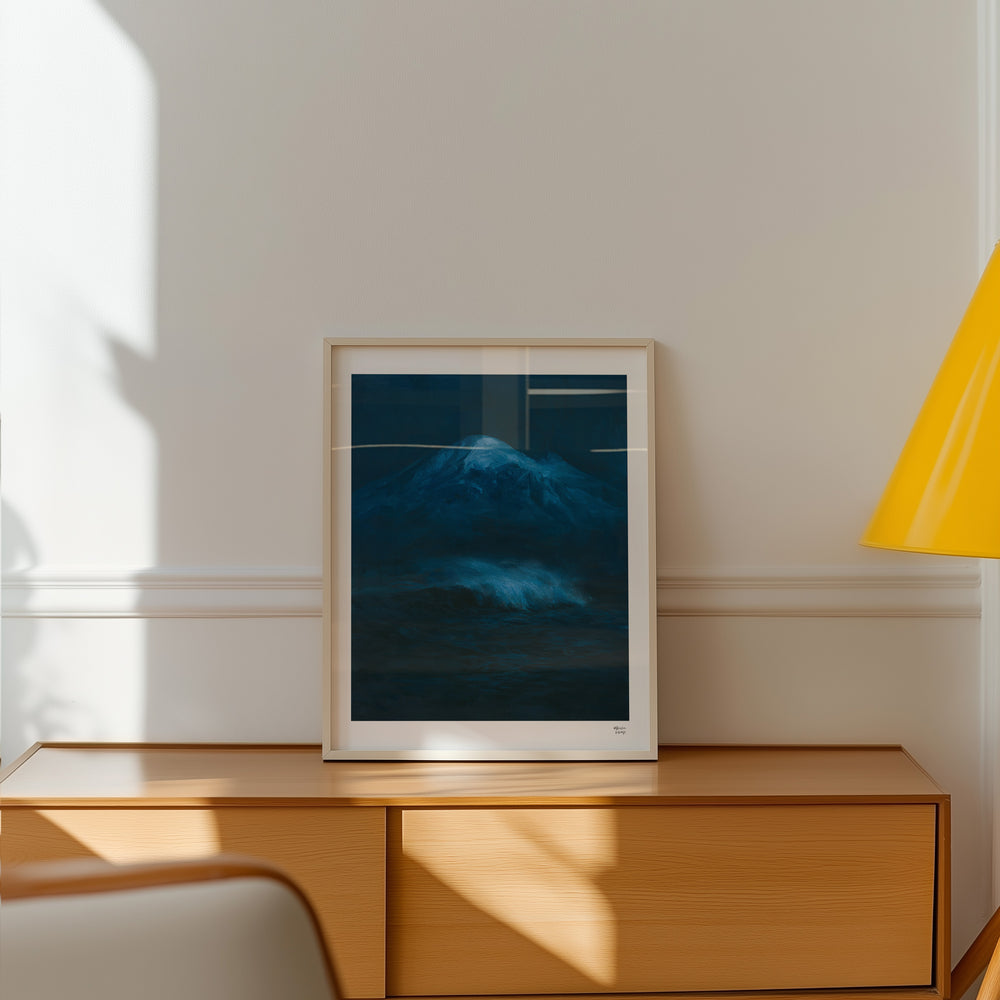 A framed art print featuring eaturing a dark blue painting of a snow-capped Mount Baker mountain above a churning ocean wave, under a light-colored frame, styled on a wooden console table against a cream wall with warm sunlight and shadows.
