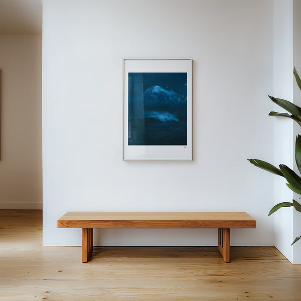 A framed art print depicting eaturing a dark blue painting of a snow-capped Mount Baker mountain above a churning ocean wave,  hangs on a white wall above a minimalist wooden bench in a bright, modern room with light wood floors and a green plant on the right.