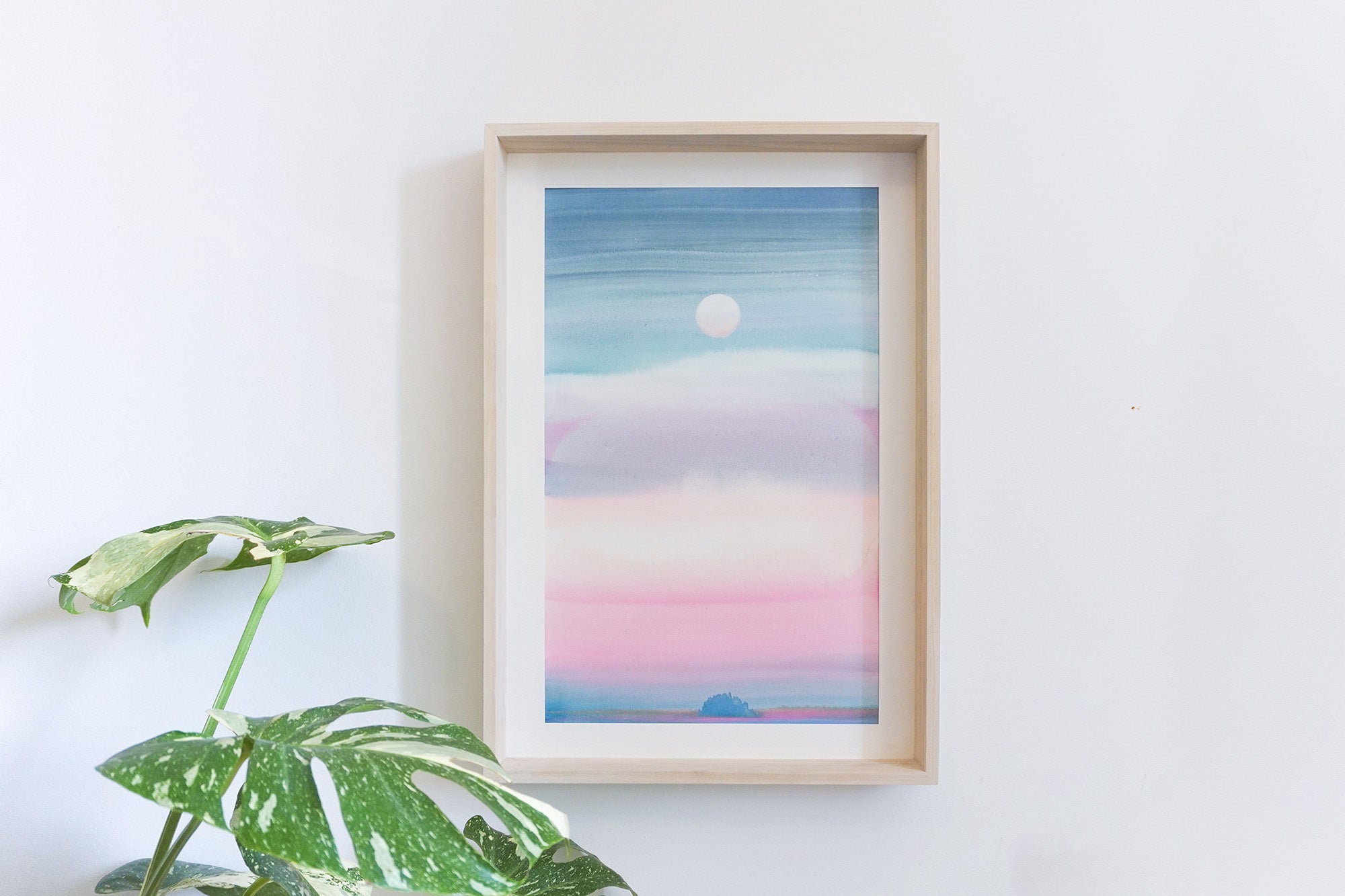 A framed pastel colour landscape watercolour featuring a blue sky, pink clouds, and a white moon, blue island displayed on a white wall next to a green plant.