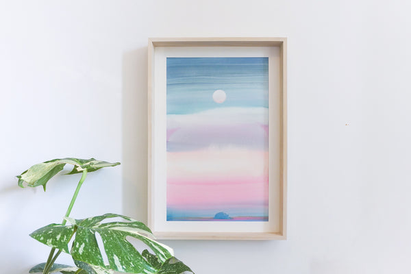 A framed pastel colour landscape watercolour featuring a blue sky, pink clouds, and a white moon, blue island displayed on a white wall next to a green plant.